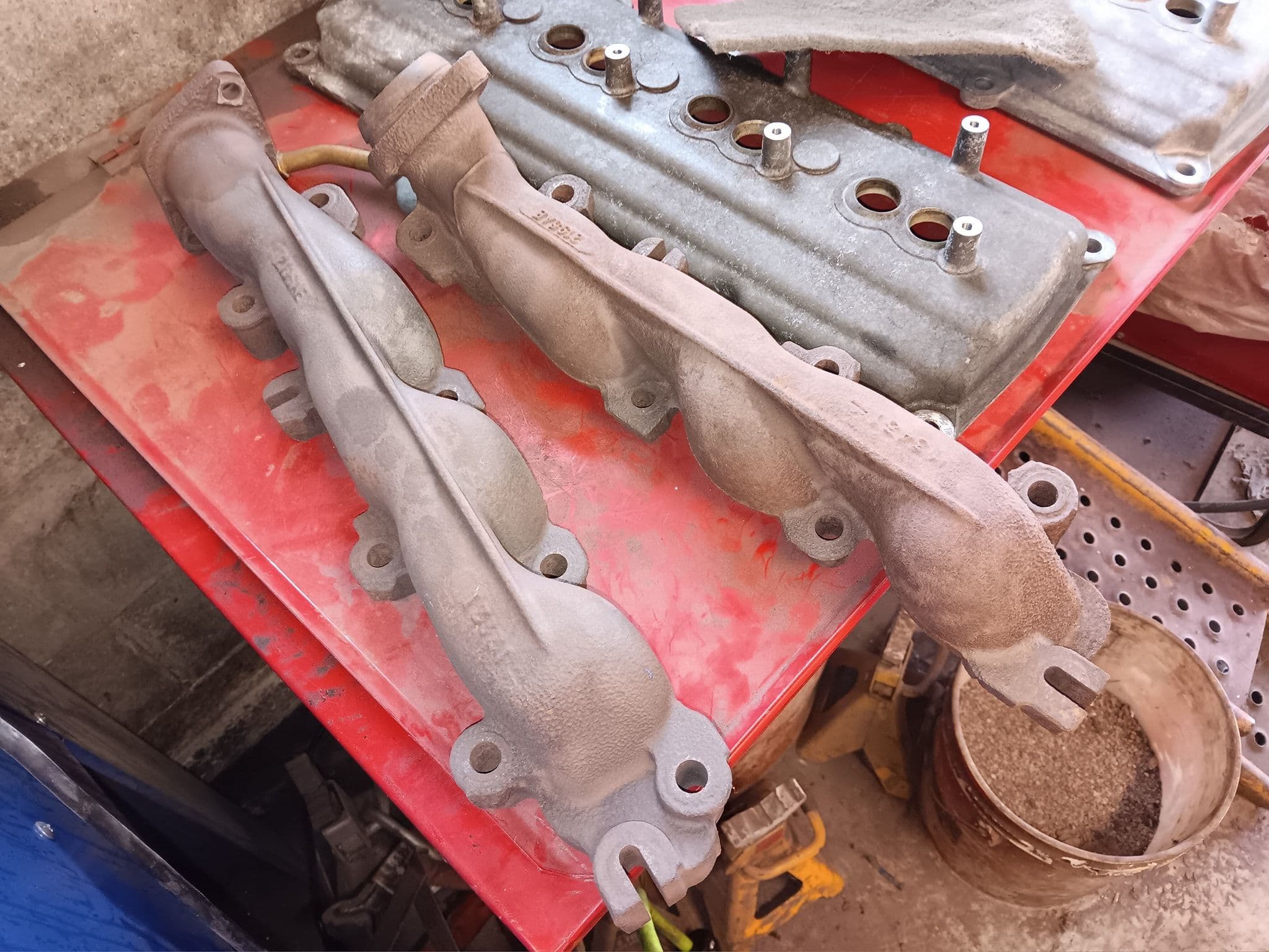 Used metal engine parts including intake manifolds on a red workbench. Dusty and worn condition.